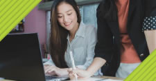 Smiling woman sitting at laptop while colleague shows work on desk with text overlay Use Your Freelance Website to Land Writing Clients