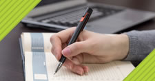 photo of a woman's hand writing in a notebook on a desk with a laptop nearby