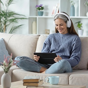 Woman sitting comfortably on a couch wearing headphones, smiling while using a tablet, with books and a coffee mug nearby, showing learning at home.