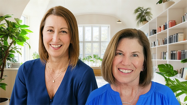 Rebecca Matter and Katie Yeakle standing together in a bright room with large windows, bookshelves, and indoor plants in the background.